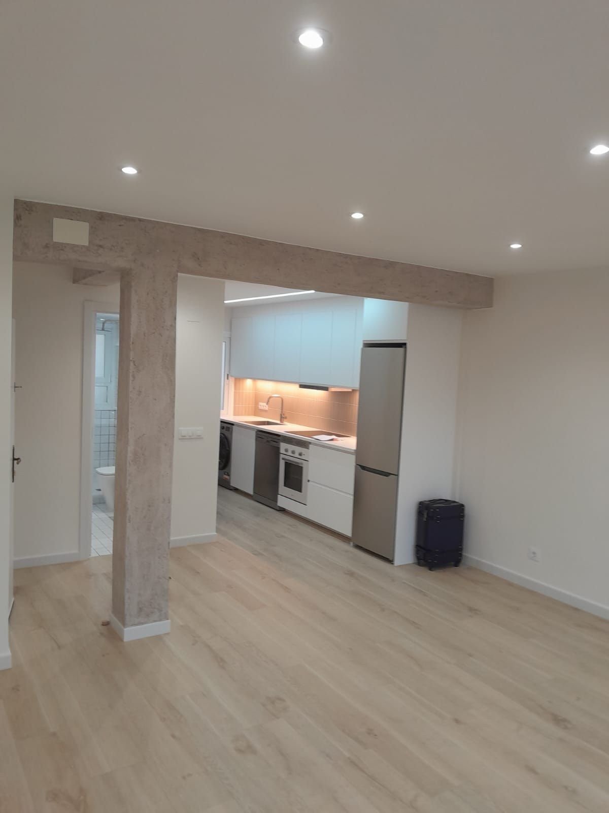 Salón-cocina abierto con viga y pilar de madera, cocina blanca y suelo claro